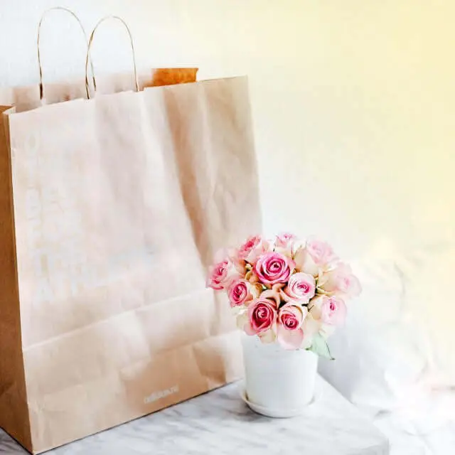 Paper shopping bag standing up and small vase of pink flowers sitting on a marble table against a yellow wall