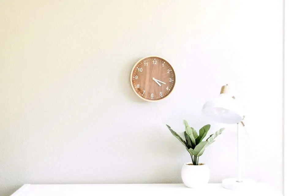 Simple desk set up with a wooden clock, small white vase with a green fern, and white lamp against a yellow wall