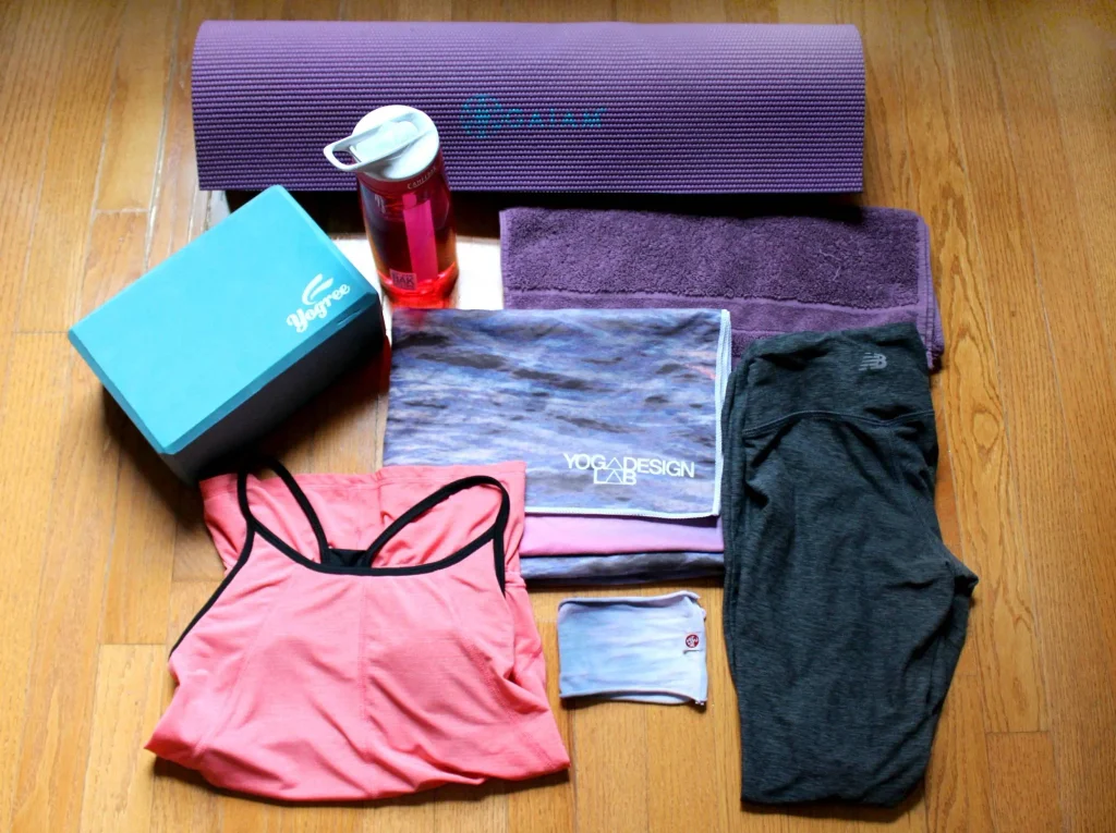 What to bring to a hot yoga class: purple mat, water bottle, yoga block, towels, workout outfit, and headband laid out on a wood floor