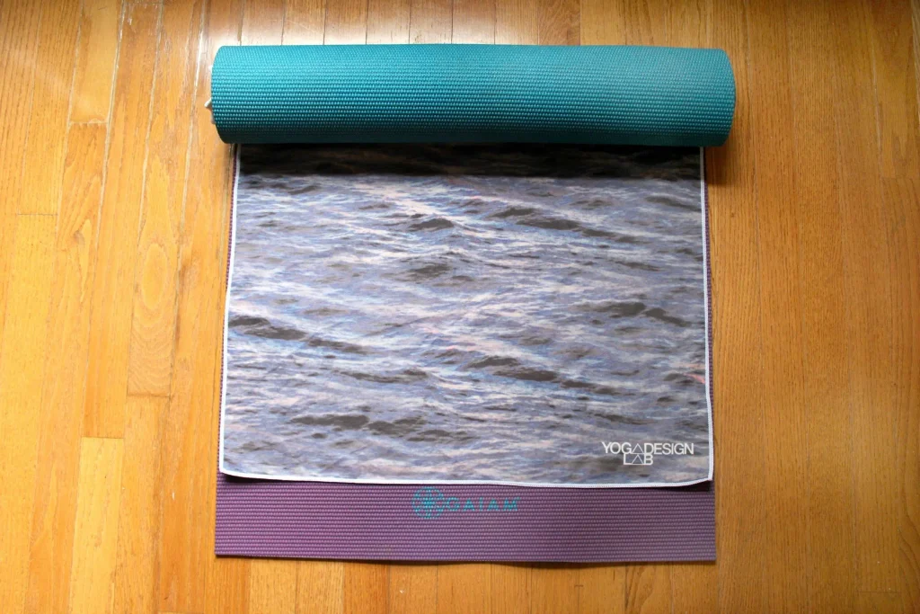 Purple Gaiam yoga mat partially covered by a Yoga Design Lab towel with a rippling water print, with a teal mat rolled at the top