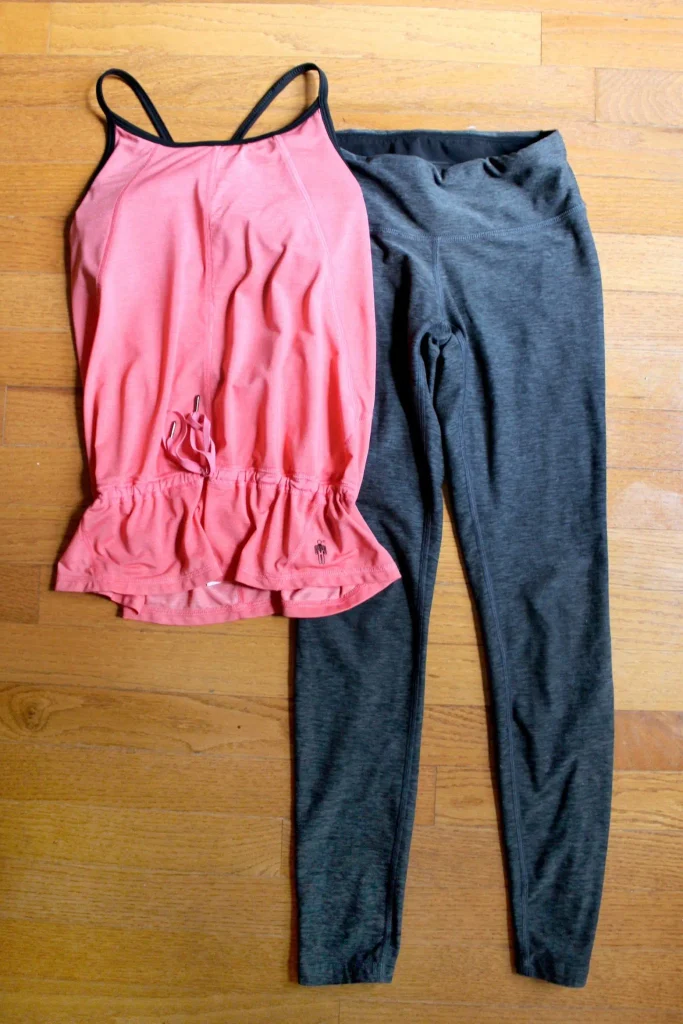 Coral racerback workout tank with a ruched hem and dark gray yoga leggings laid flat